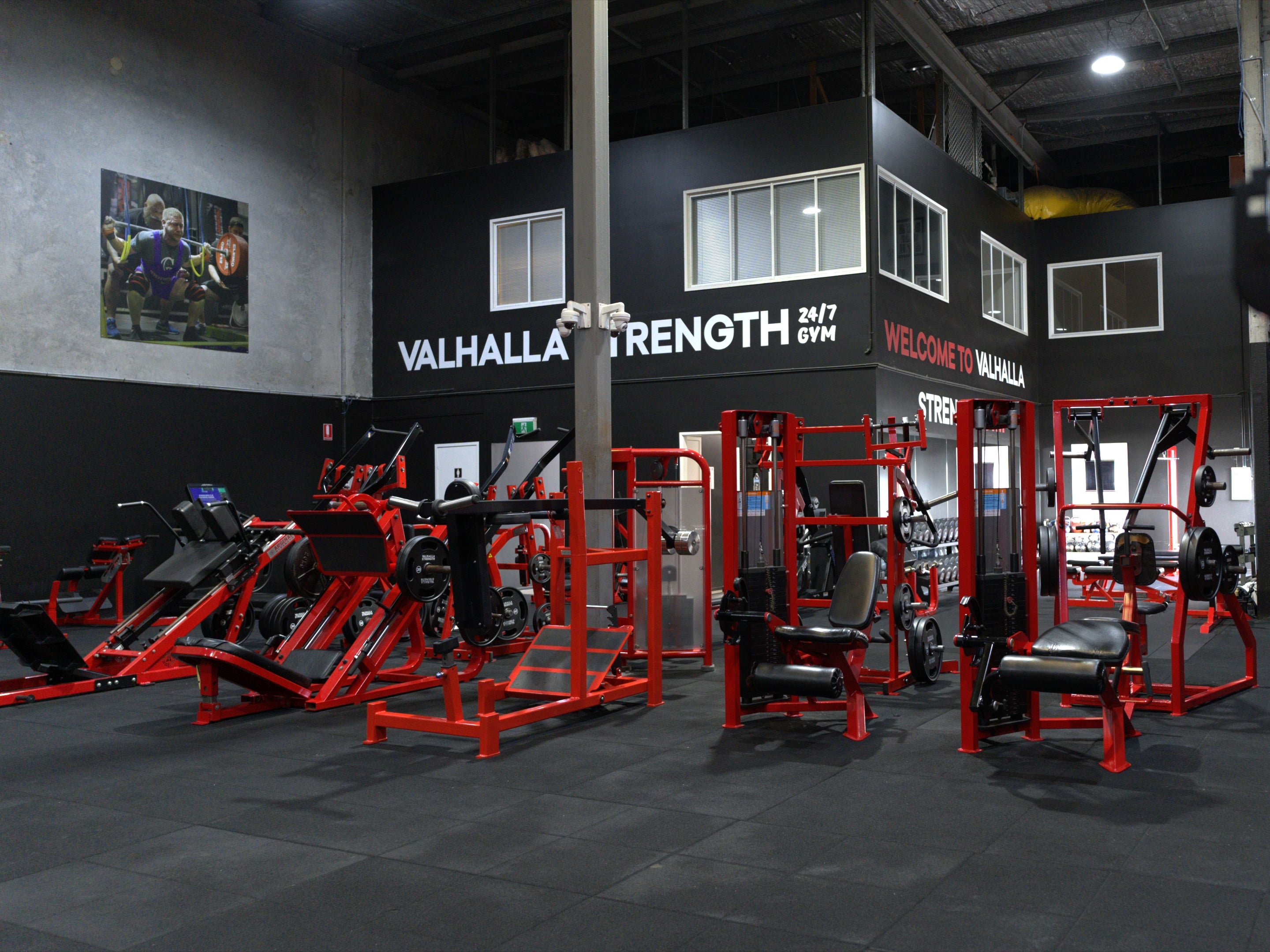 A gym interior with multiple red and black strength training machines arranged in a large black-and-gray facility. Visible wall signage reads 'VALHALLA STRENGTH 24/7 GYM' and 'WELCOME TO VALHALLA'.